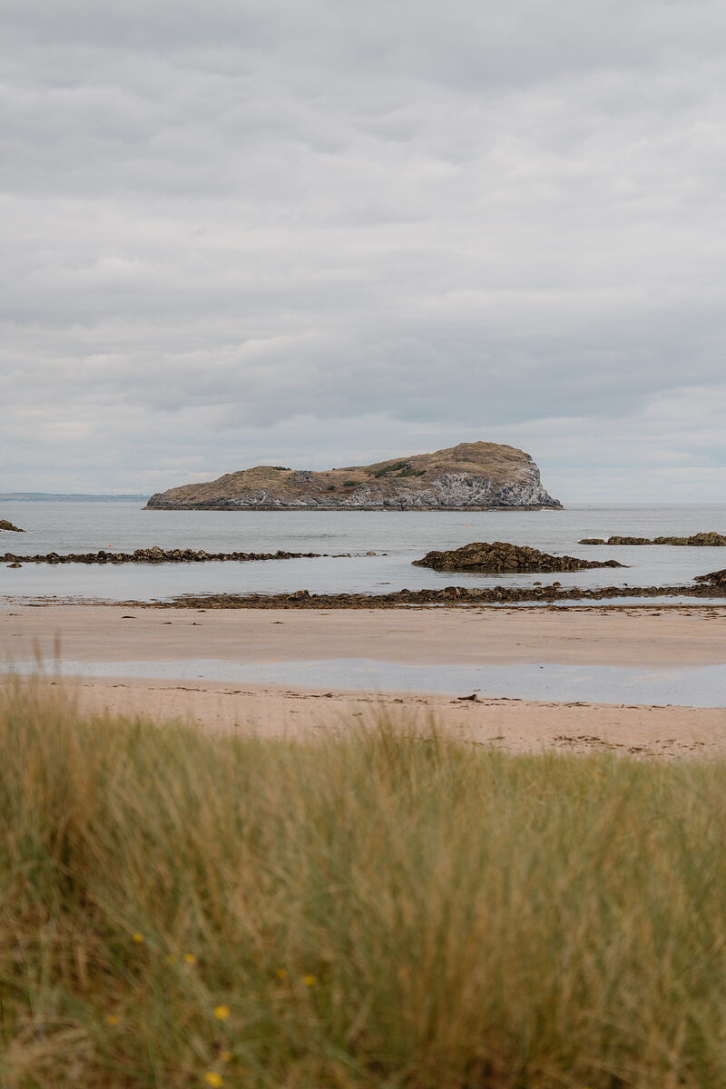 A rocky island rising from the sea beyond a sandy beach and low tide pools.