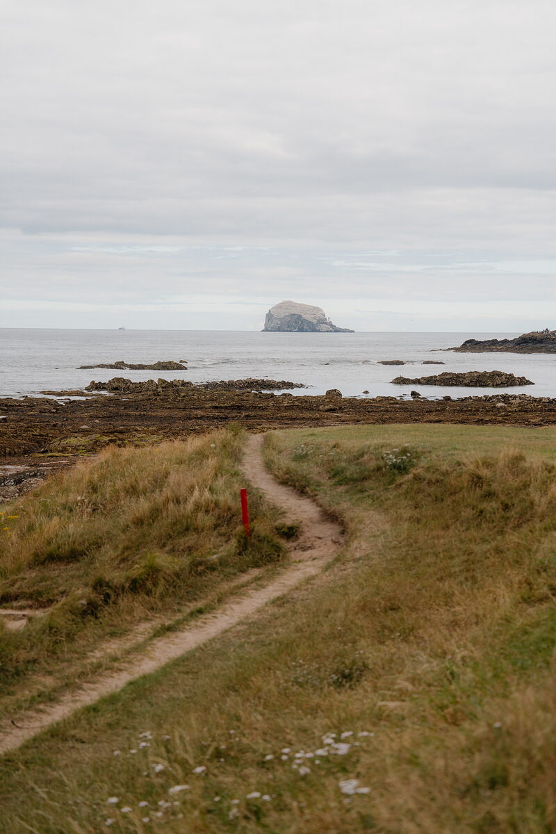 A rocky island rising from the sea beyond a coastal path, a marker post standing in the grass.