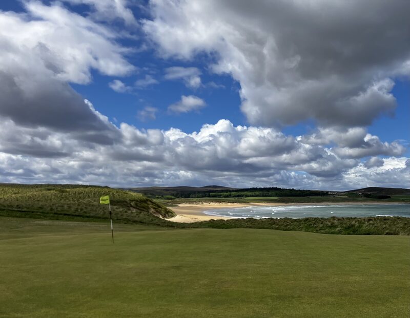 A green with a yellow flag at Reay, with marram dunes and the sands of Sandside Bay beyond under a big sky.
