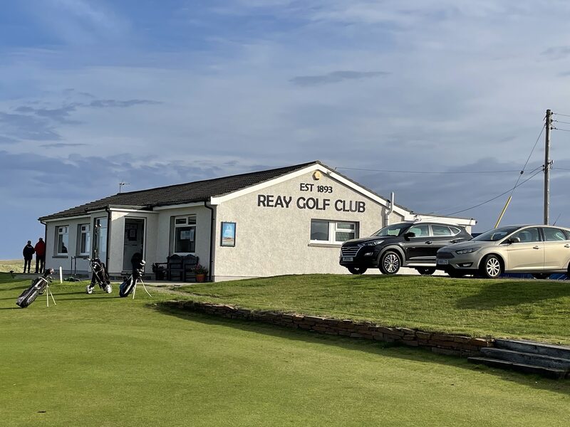 The clubhouse of Reay Golf Club, lettered Est 1893 Reay Golf Club, with golf bags on trolleys outside and cars in the car park.