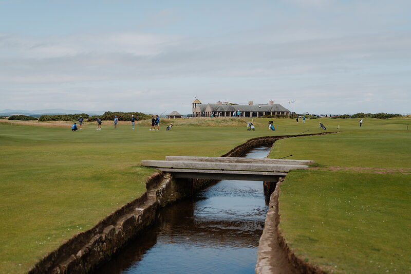 Golfers crossing the Swilcan Bridge at St Andrews Links, the clubhouse standing beyond the green.