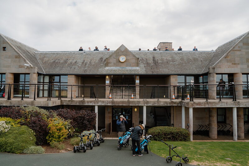 St Andrews Links' clubhouse, spectators watching from its rooftop terrace above golfers with trolleys.
