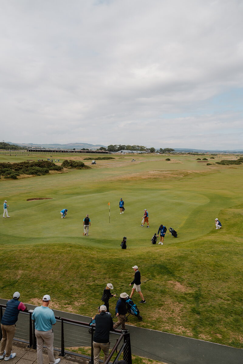 Golfers putt out on a green while spectators watch from a railing, grandstands and marquees beyond.