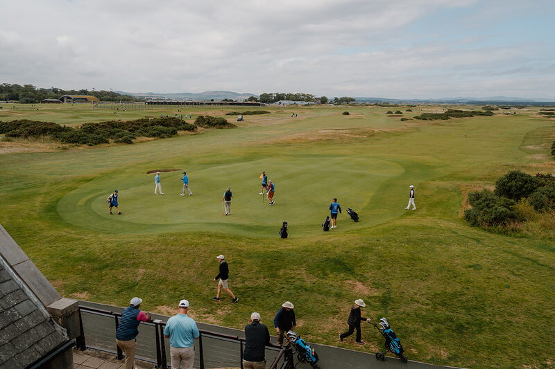 Golfers gather on a green as spectators look on from a raised platform.