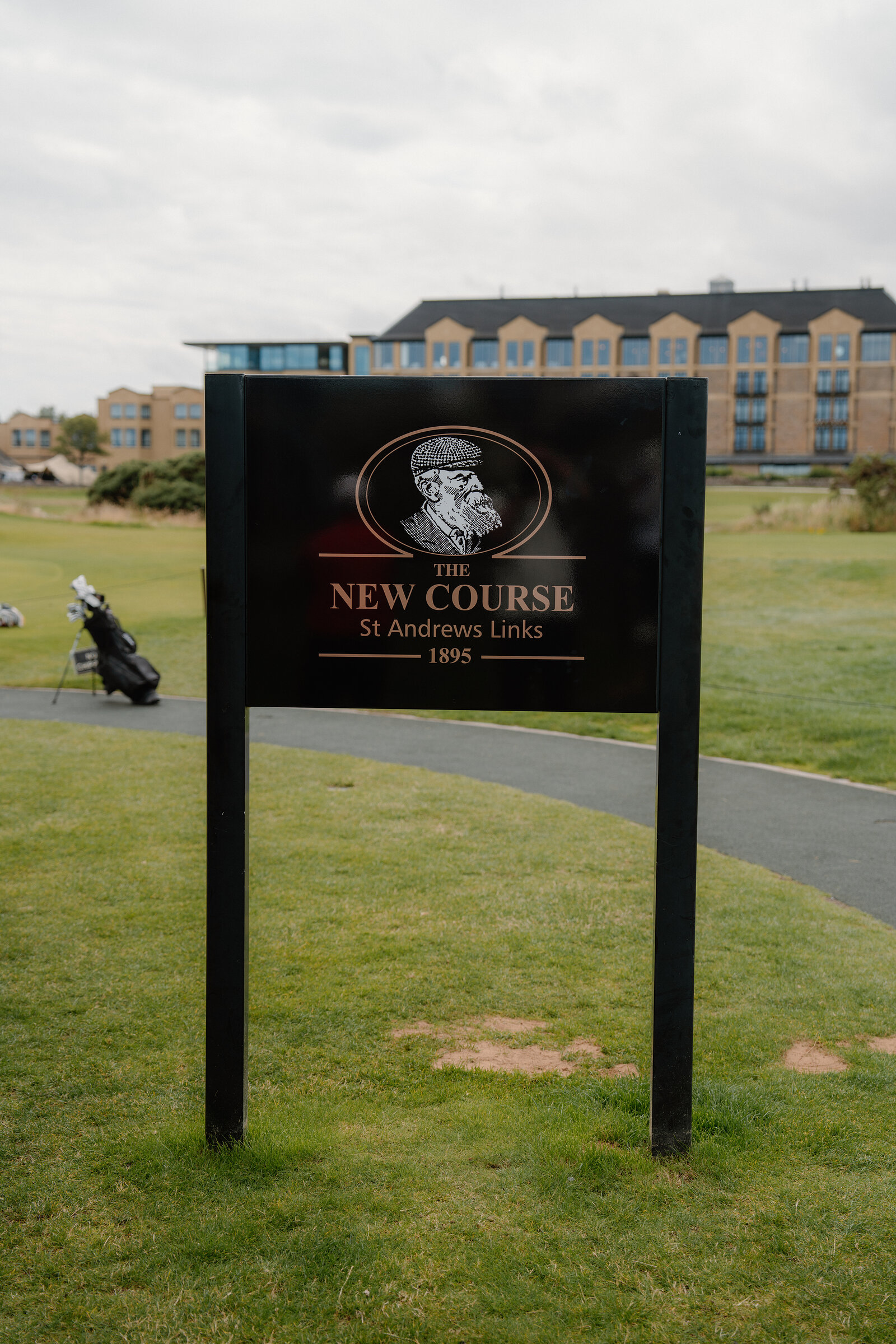 St Andrews Links' own sign for the New Course, dated 1895, stands before a hotel building with golf bags nearby.