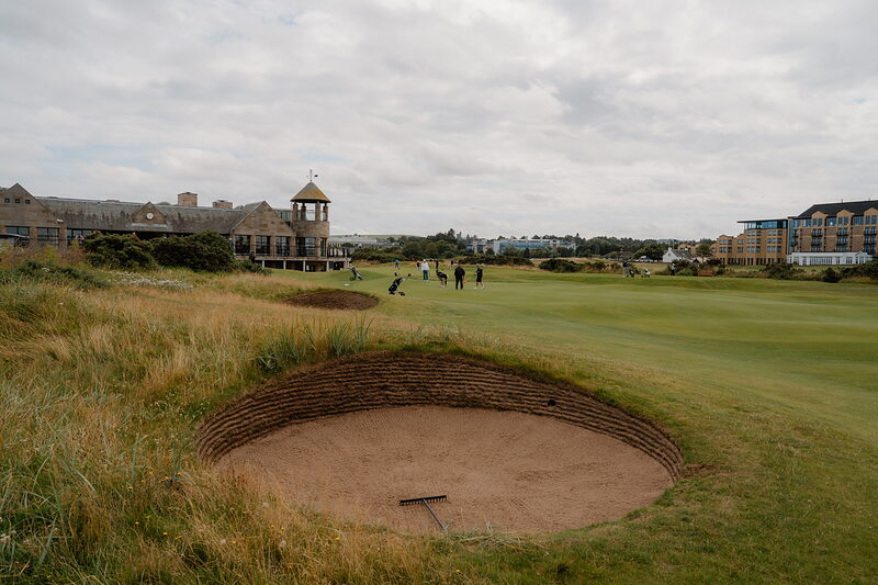 Golfers putt out on a green backed by a clubhouse tower, a bunker curving in the foreground.