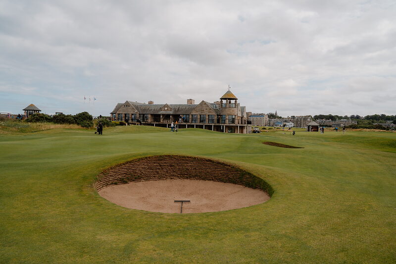 Golfers cross a green backed by a timber-clad clubhouse and tower, a bunker in the foreground.