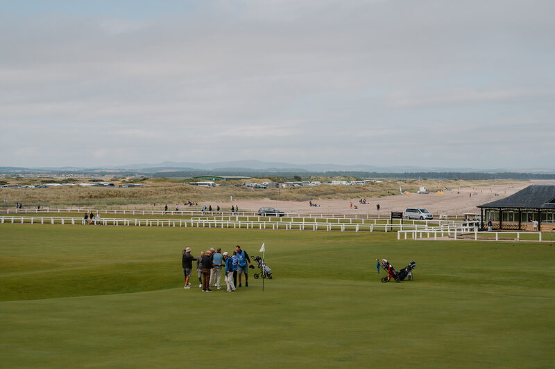 Golfers gather on a green with a sandy beach and grandstand stretching away behind them.