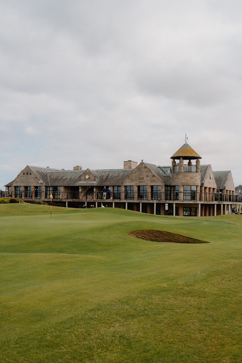 A clubhouse tower rises behind golfers on the fairway, a bunker curving in the foreground.