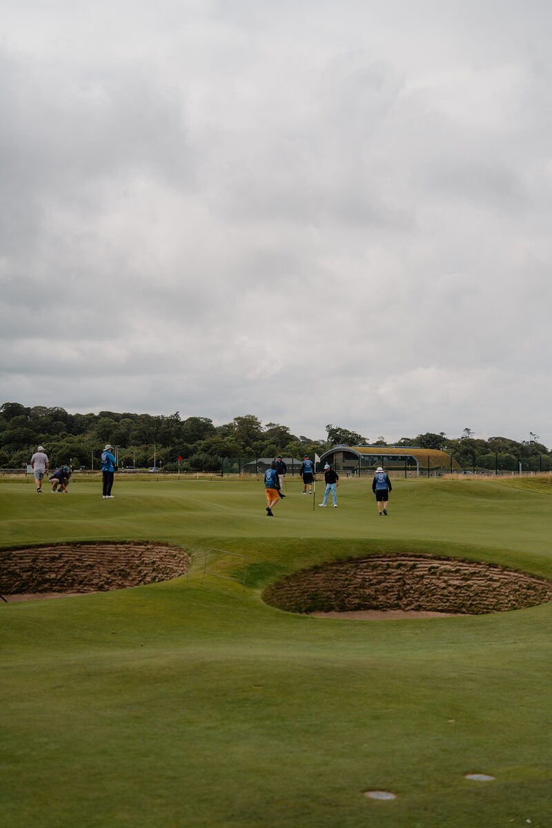 Golfers cross a green between two bunkers, buildings and trees lining the fairway beyond.