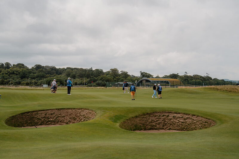 Golfers cross a green between two bunkers, a marquee and trees lining the fairway beyond.