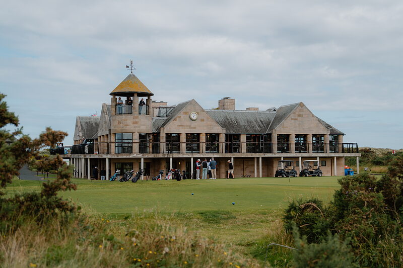 A clubhouse tower rises behind golf bags and golfers gathered on the fairway.