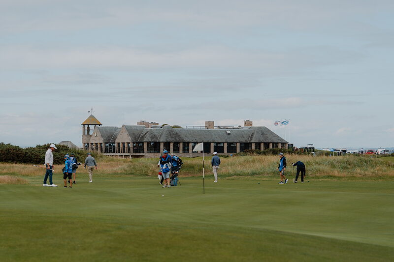 Golfers putt out on a green as a clubhouse and flags stand behind them.