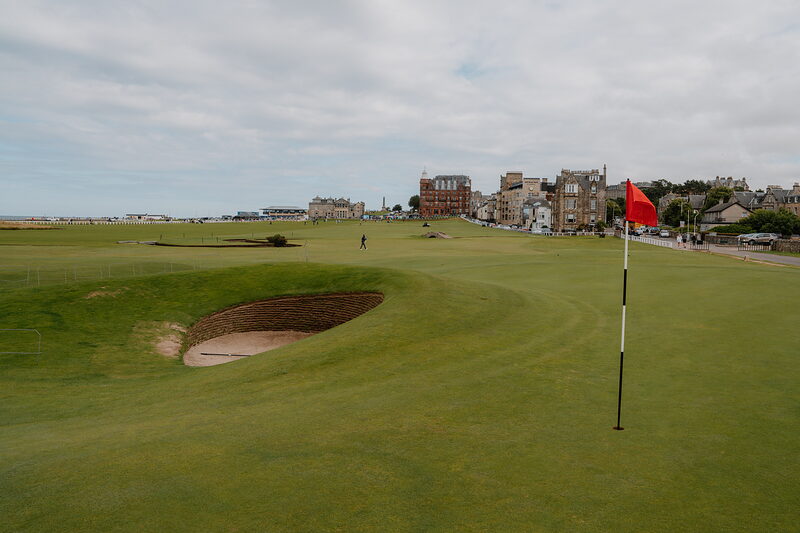 A bunker guards a green flying a red flag, the town of St Andrews rising beyond.