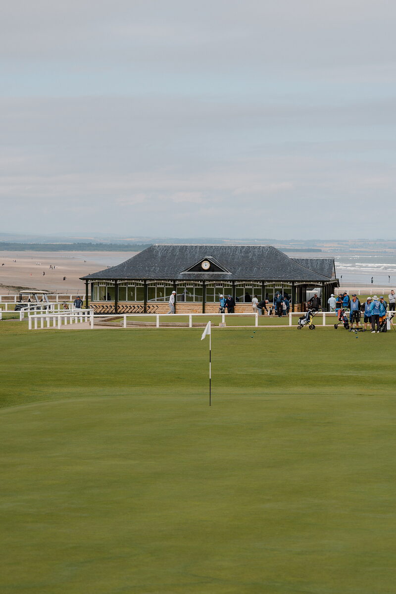 A clubhouse pavilion overlooks a beach as golfers gather near a green flag.