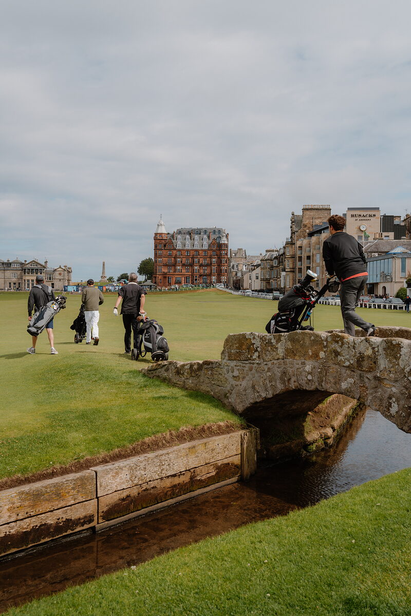 Golfers wheel their clubs across the Swilcan Bridge as the town rises behind them.