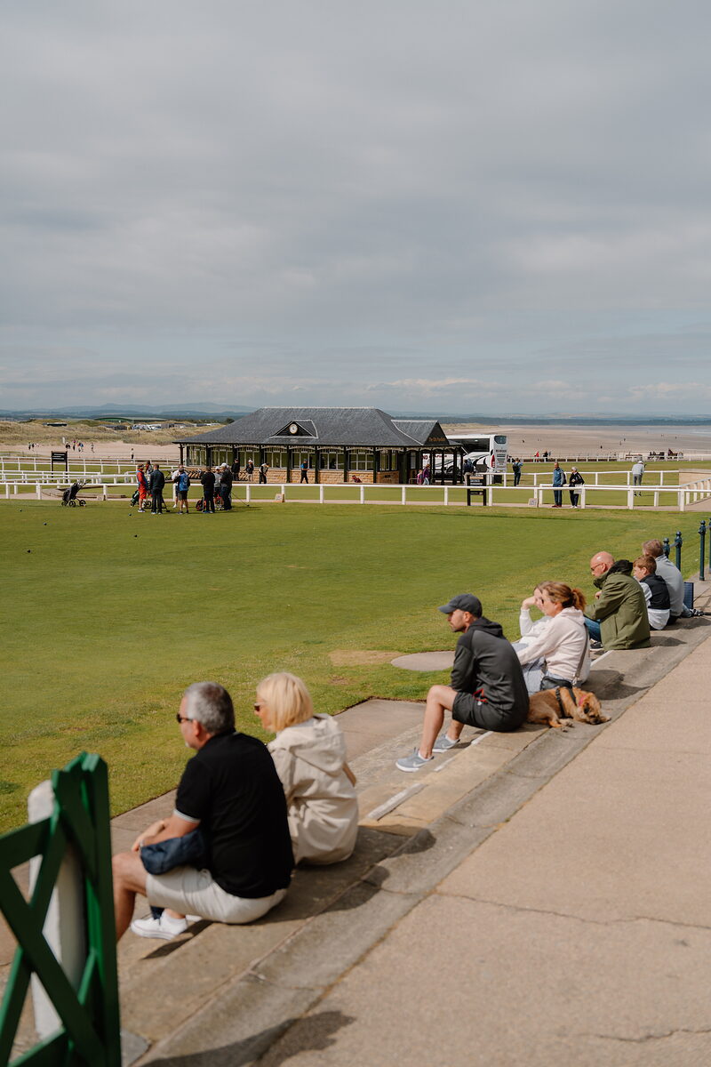 Spectators sit along a rail watching golfers gather near a clubhouse pavilion above West Sands beach.