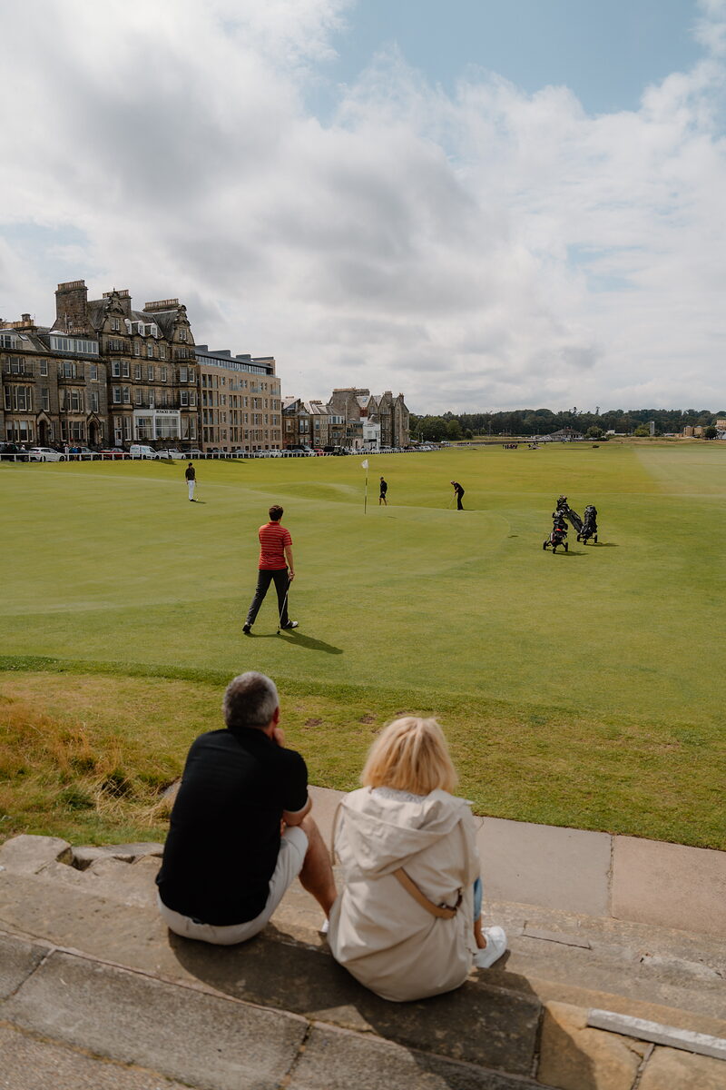 Spectators sit on a low wall watching golfers putt on a green before the Old Course hotels.