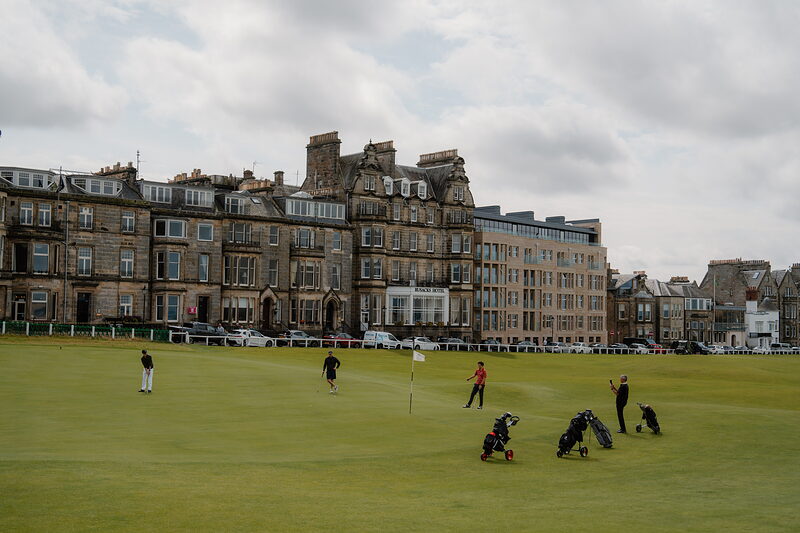 Golfers gather on a green as hotel buildings rise along the Old Course's edge.