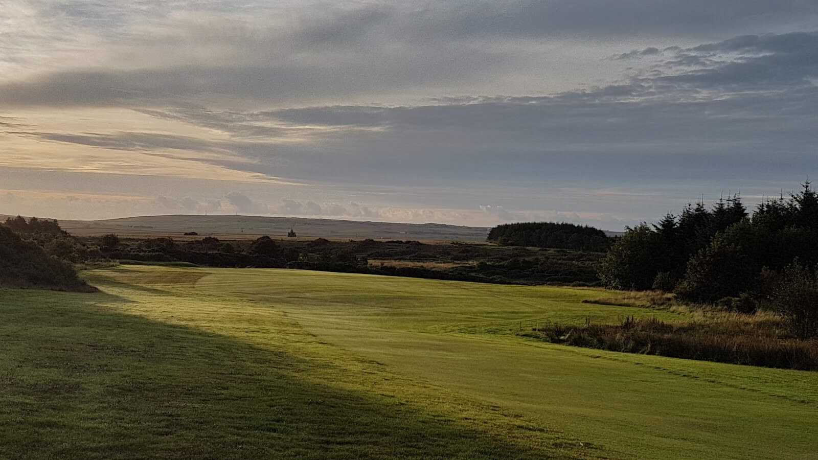 A fairway at Thurso Golf Club in low evening light, with dark conifers on the right and moorland hills on the horizon.