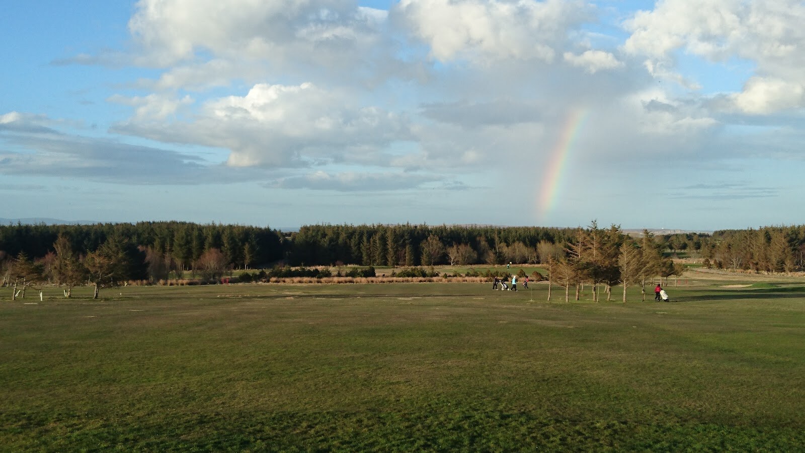 A rainbow over Thurso Golf Club, with golfers crossing wide fairways between young trees and forestry behind.