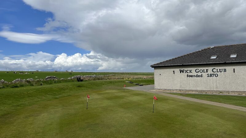 The clubhouse of Wick Golf Club, lettered Wick Golf Club founded 1870, beside a putting green with red flags.