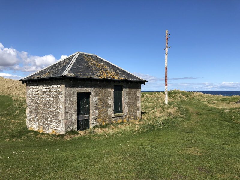 The cable house on Wick’s links, a small stone building with a slate roof, beside a red and white telegraph pole.