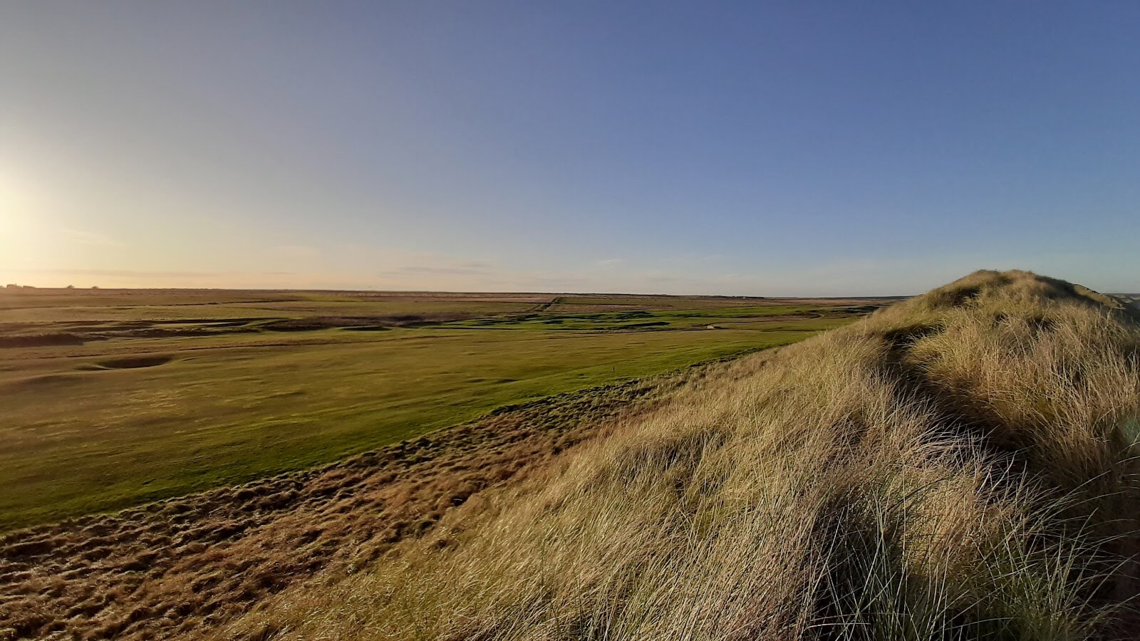 Golden marram dunes in low evening sun, looking over flat links fairways and bunkers to the horizon.
