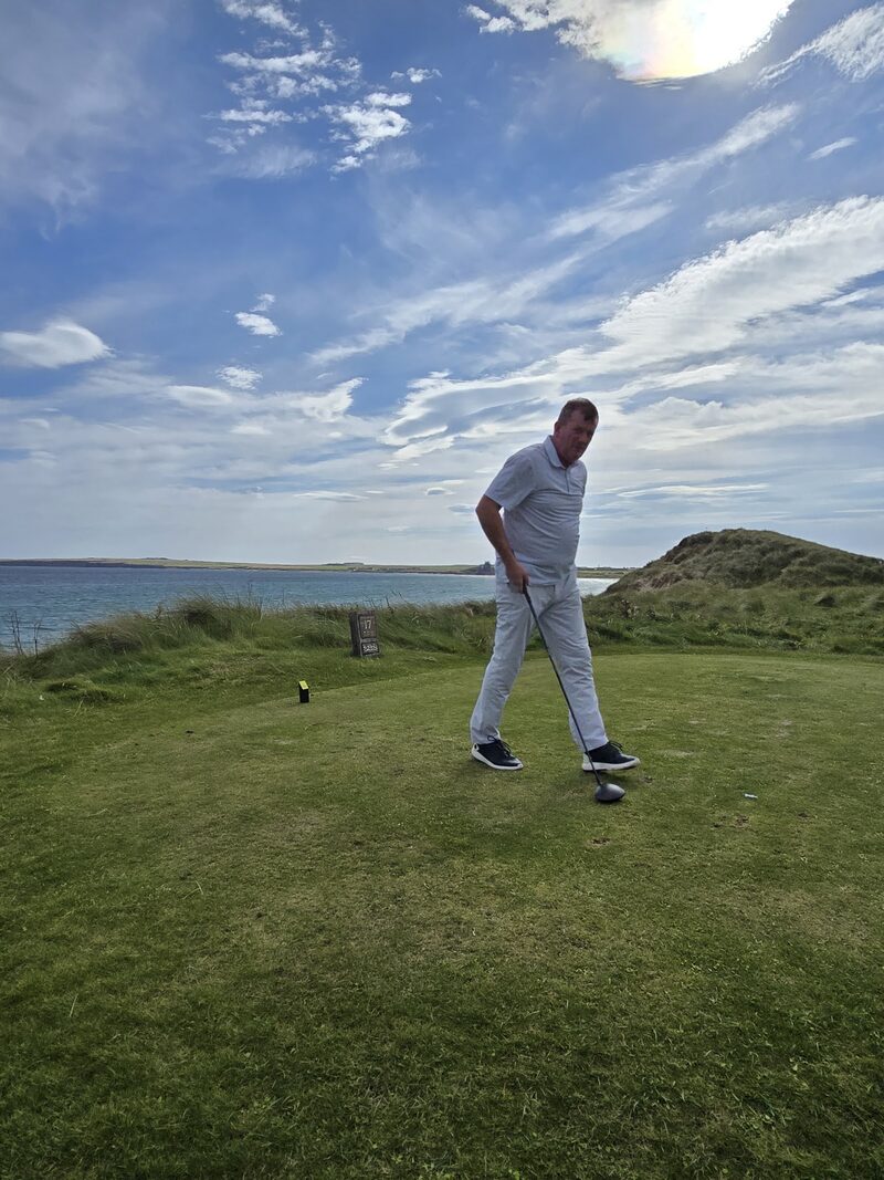 A golfer on the 17th tee at Wick, with its tee stone, the dunes and Sinclair Bay behind.
