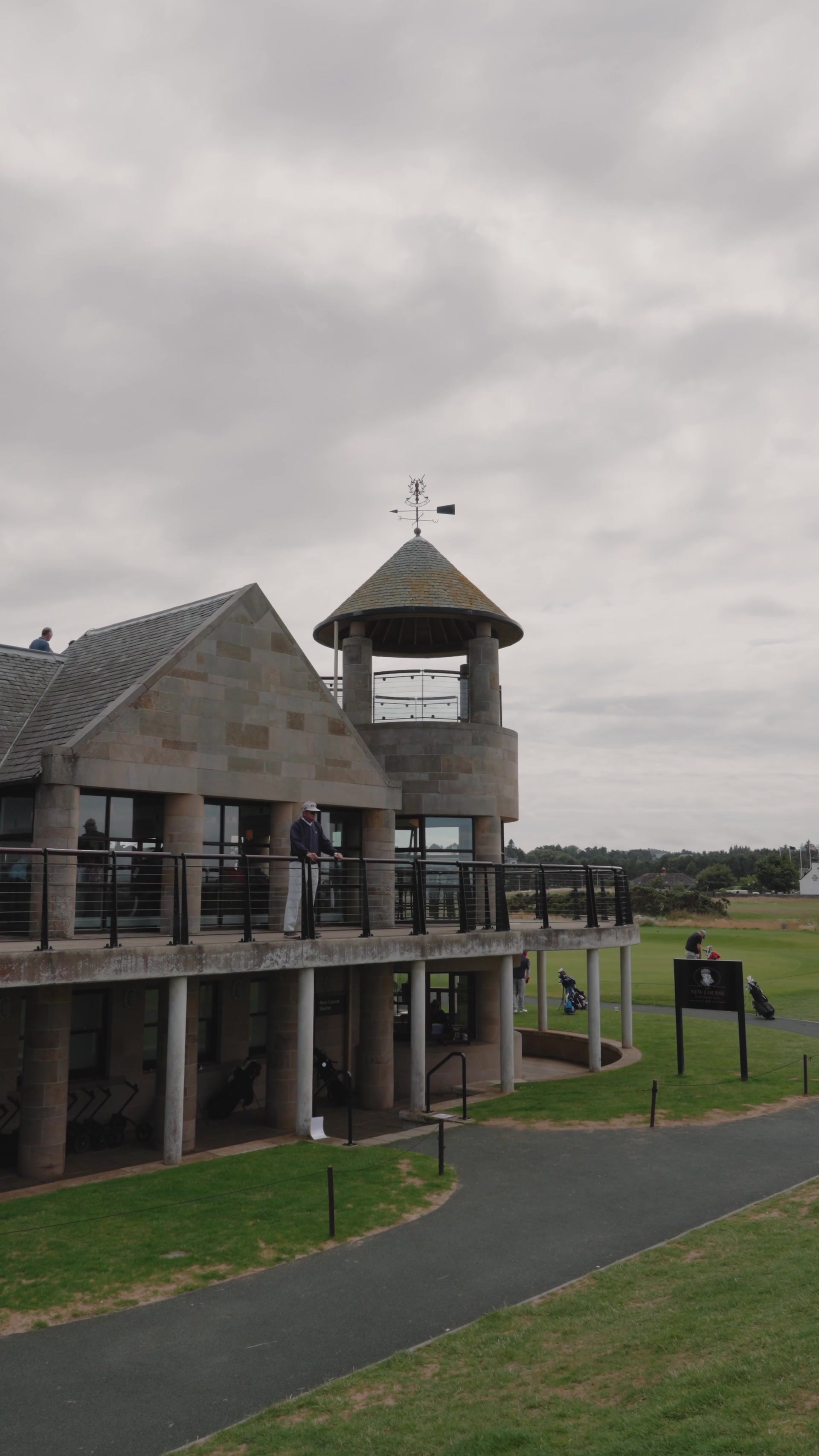 A stone clubhouse tower topped with a weathervane overlooks the New Course at St Andrews, golfers gathering on the balcony and fairway below.