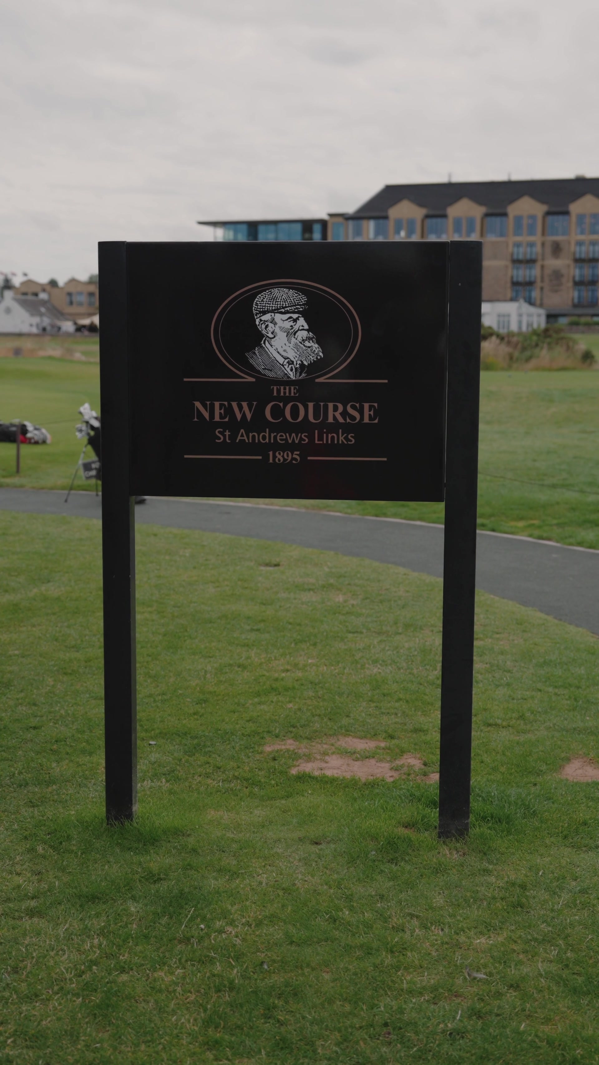 A black sign reading "The New Course, St Andrews Links, 1895" and illustrated with a flat-capped golfer's portrait stands on the grass, a sandstone hotel rising behind.