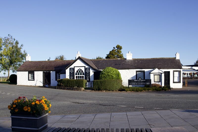 The whitewashed Famous Blacksmiths Shop at Gretna Green under a clear blue sky, with flowers in a planter by the road.