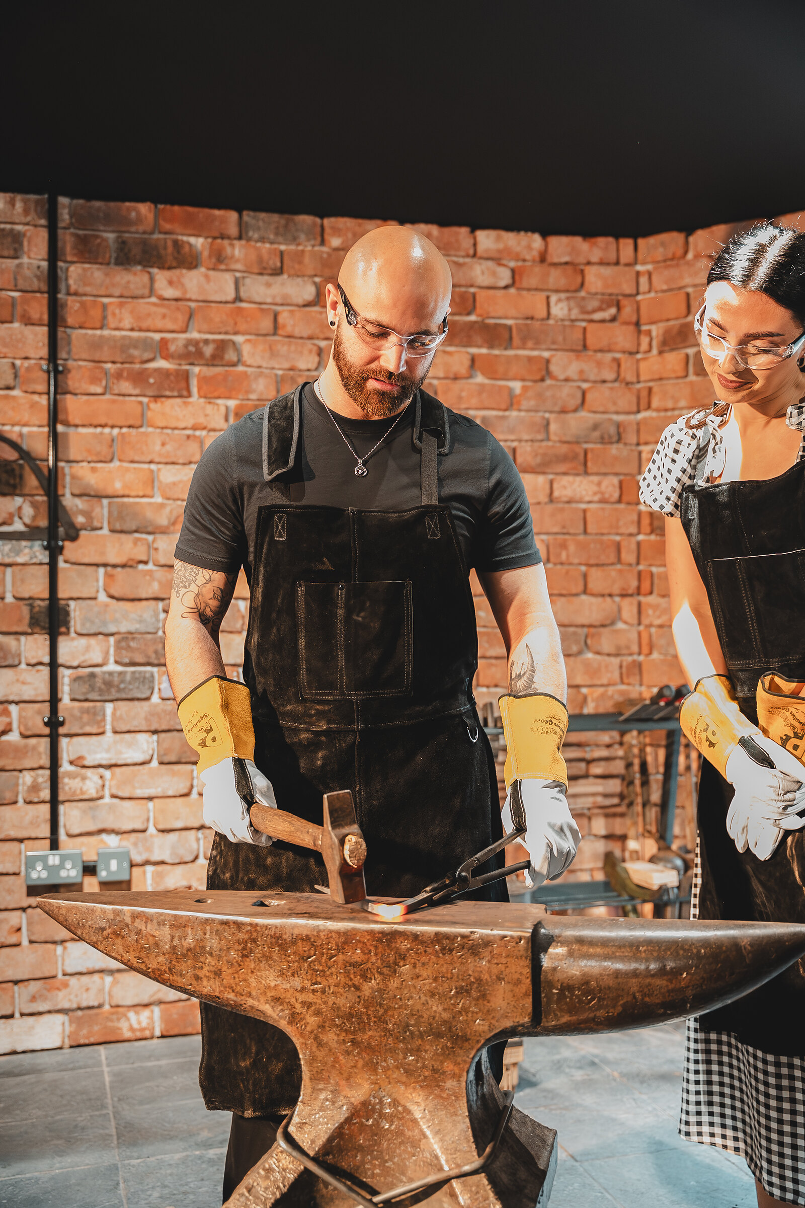 A man in a leather apron hammers at the anvil while his partner looks on.