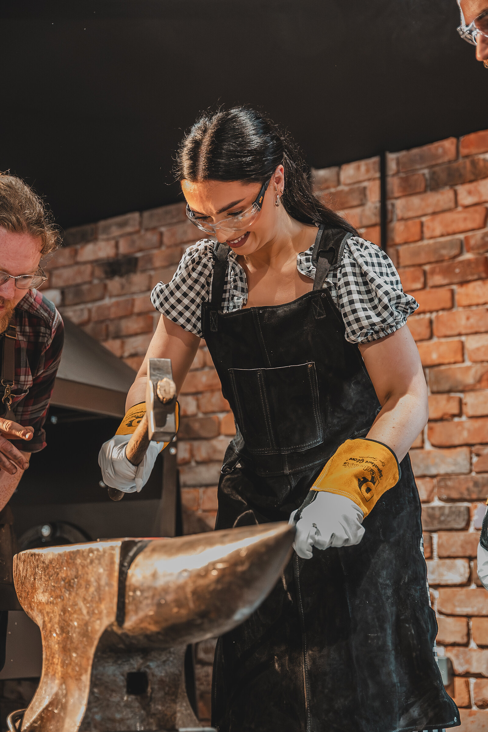 A woman in safety glasses and gloves takes her turn with the hammer at the anvil.