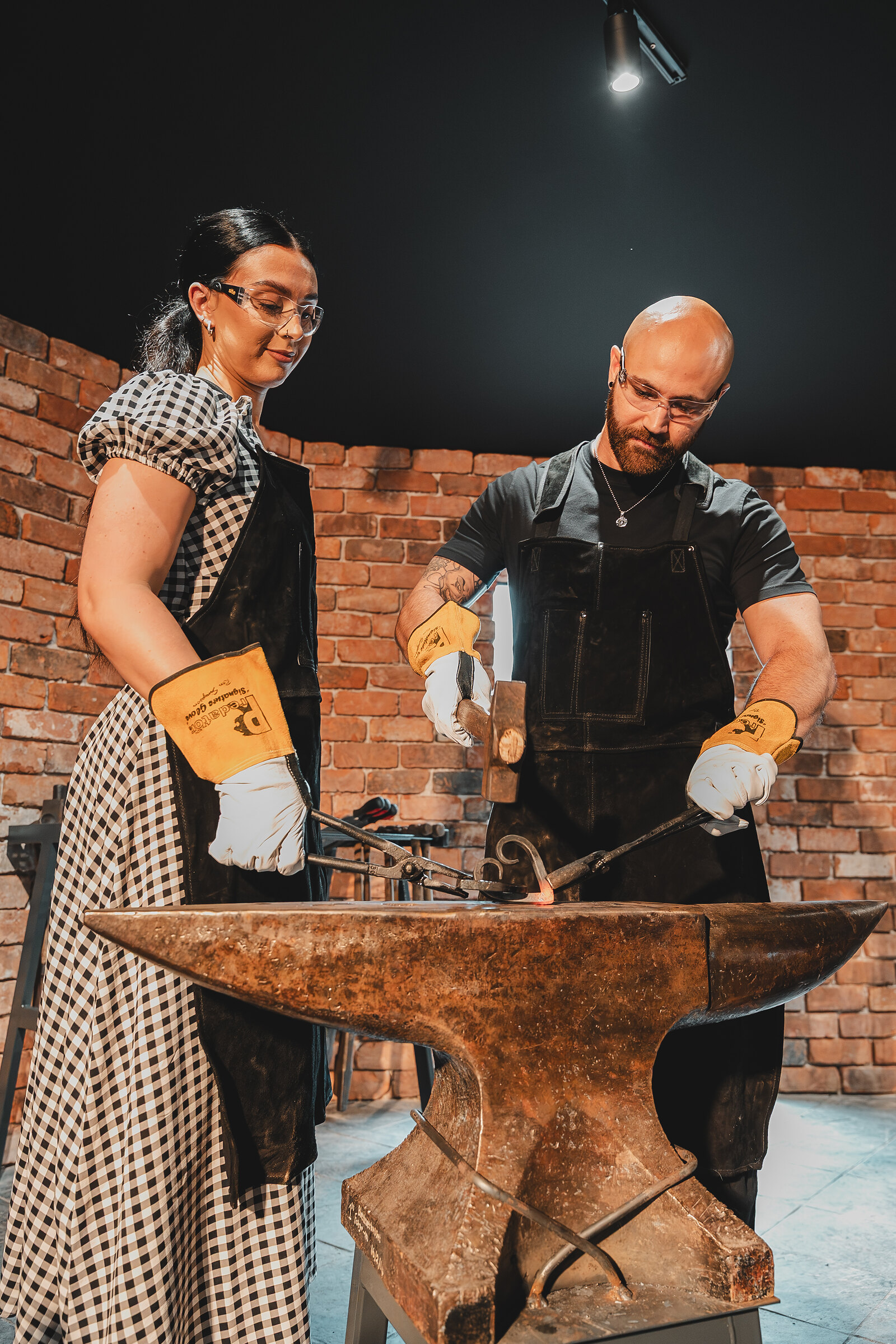 A couple stand at the anvil together, hammers in hand, under the Forge's lights.