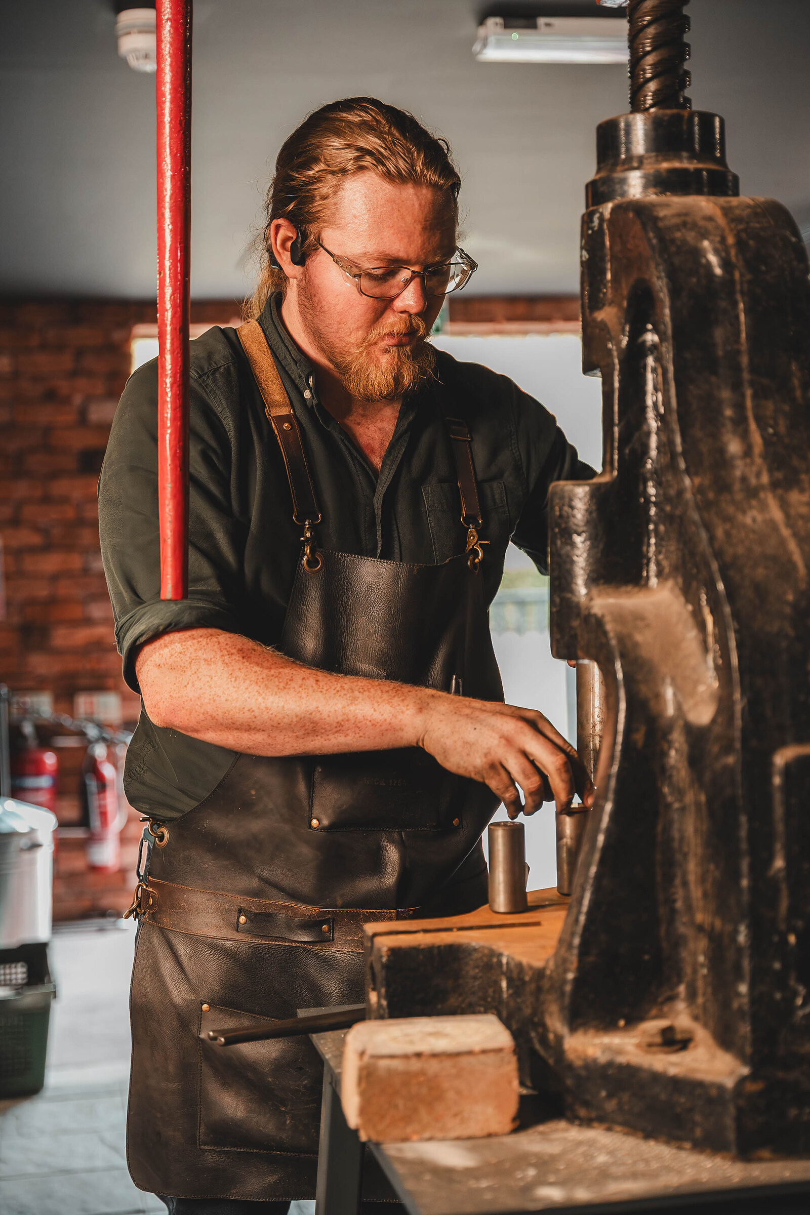The blacksmith, in a leather apron, sets up the coin press against the Forge's brick wall.