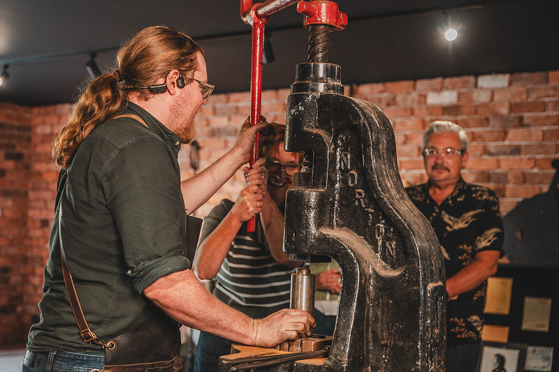 The blacksmith works the big iron coin press at the Gretna Green Forge while a visitor in a striped jumper looks on.