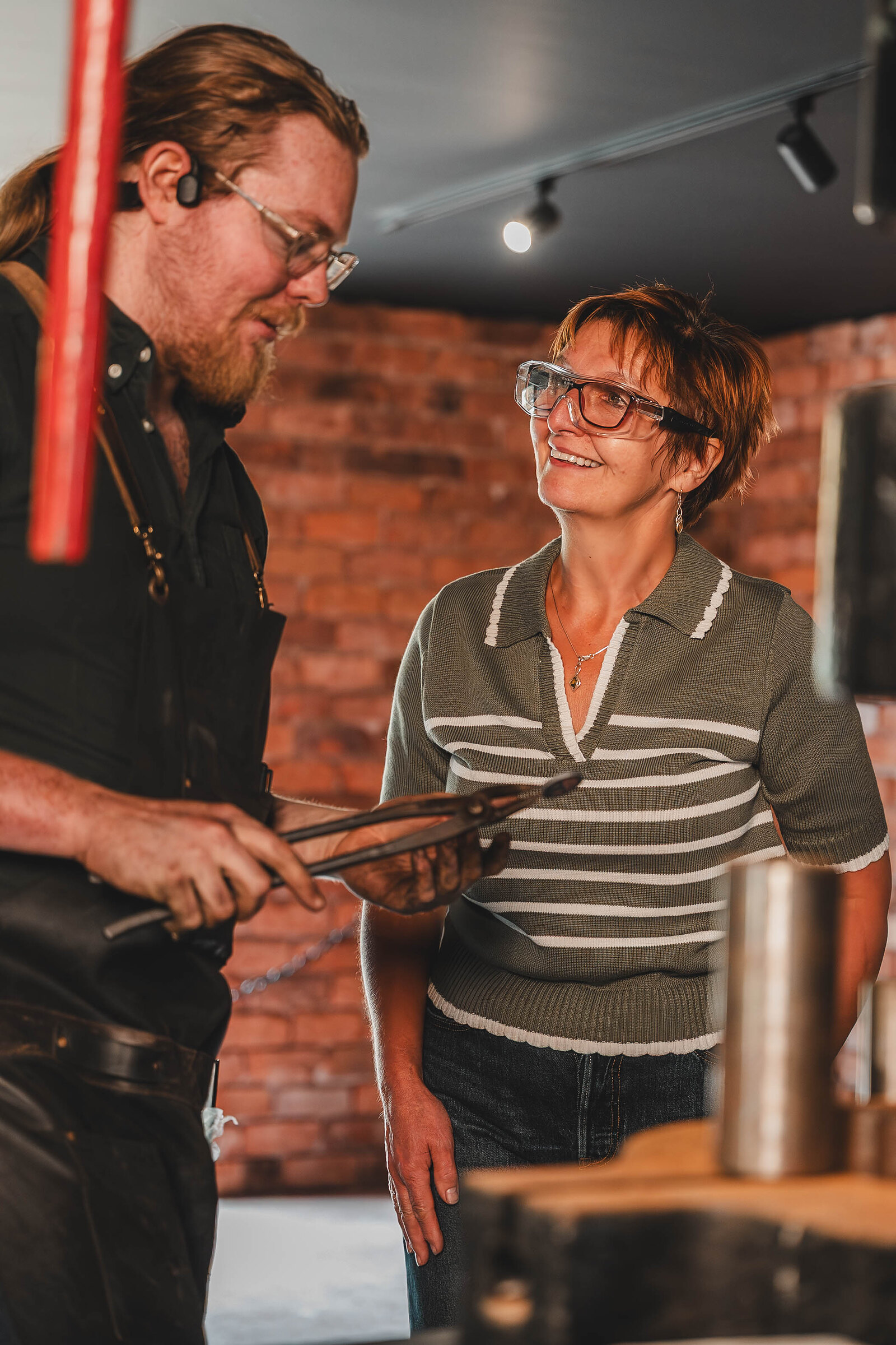 The blacksmith holds a coin in his tongs as a smiling visitor watches.