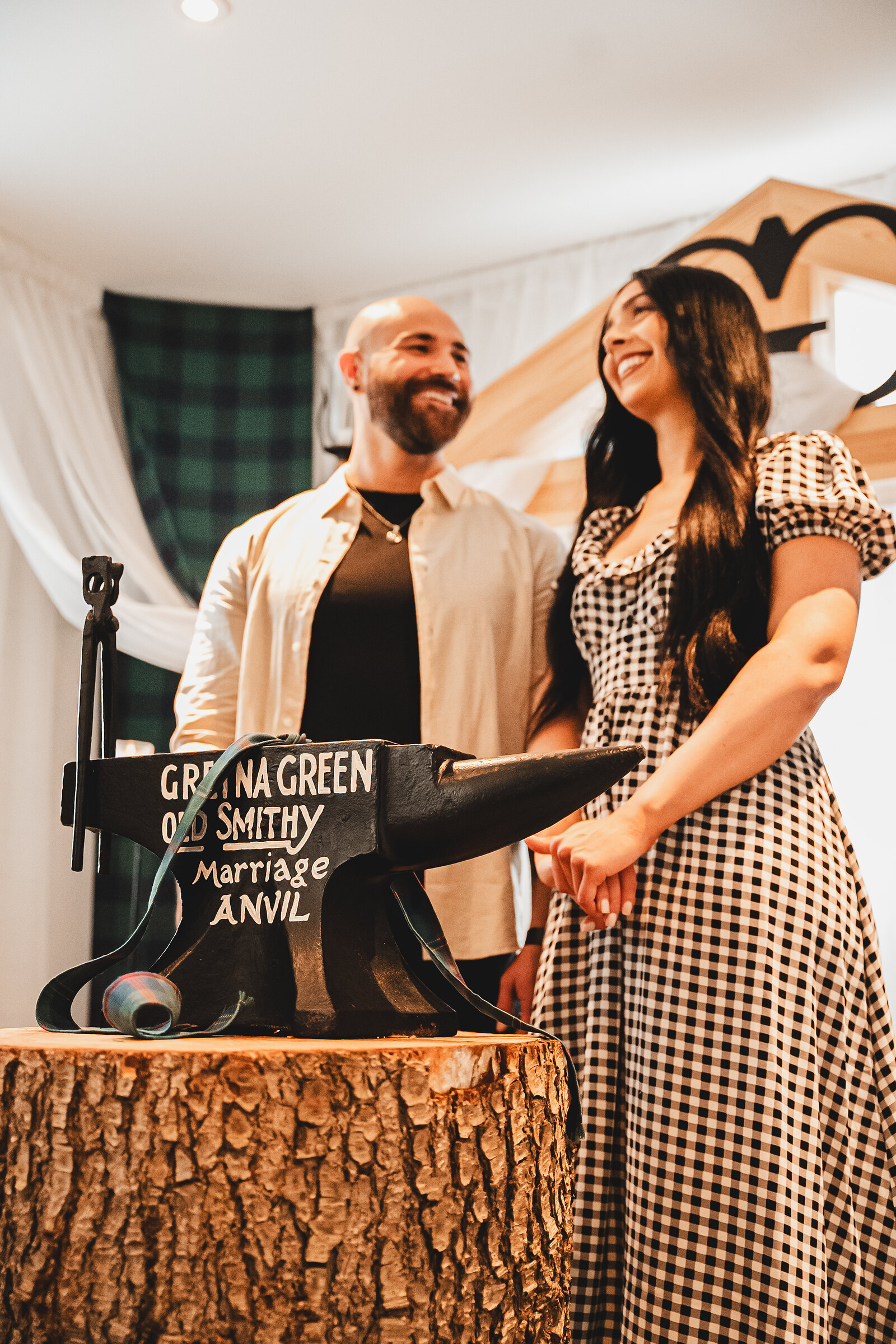 A laughing couple stand hand in hand beside the Gretna Green Old Smithy Marriage Anvil.