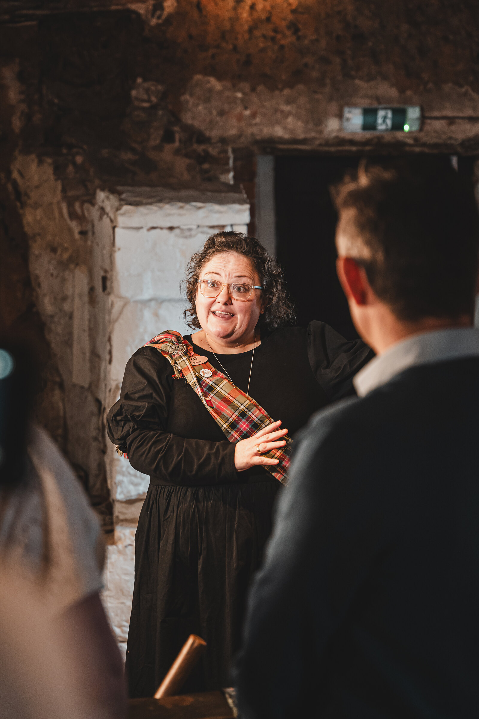 A guide in a black dress and tartan sash smiles as she begins a handfasting in the museum.