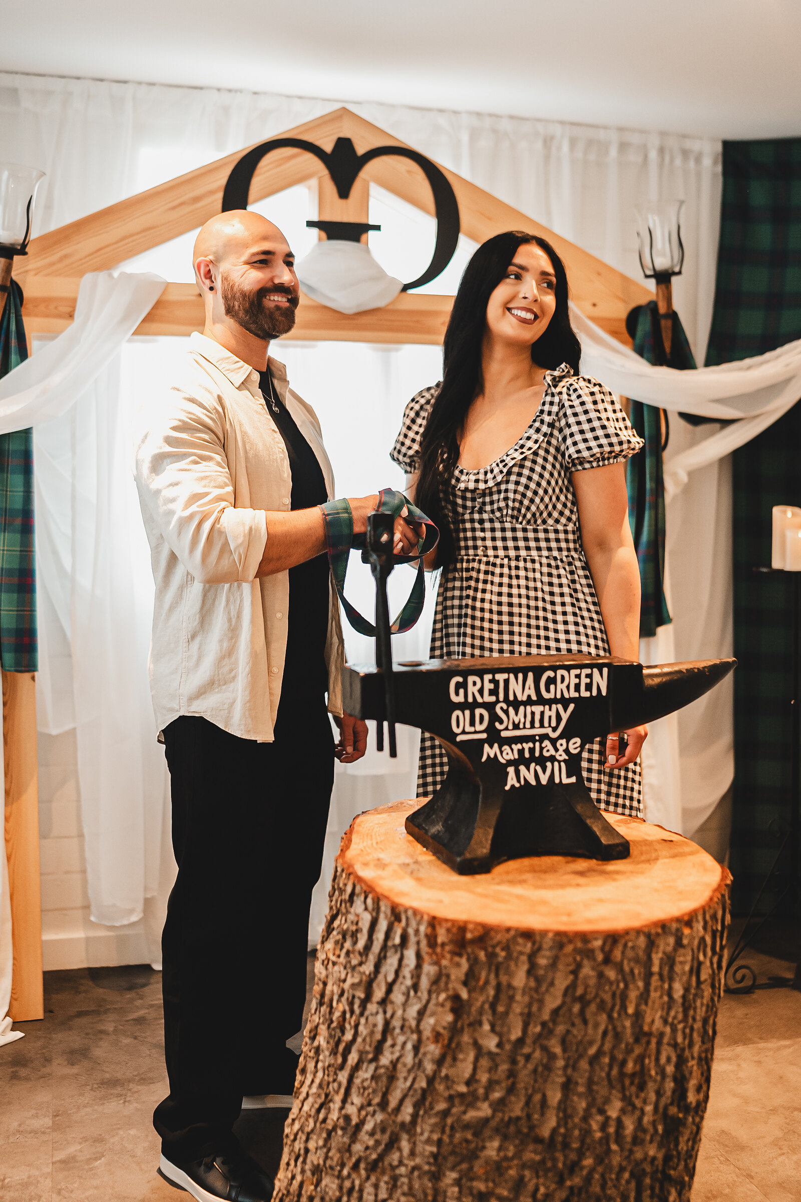 A smiling couple stand at the marriage anvil with their hands tied in tartan, under a carved heart arch.