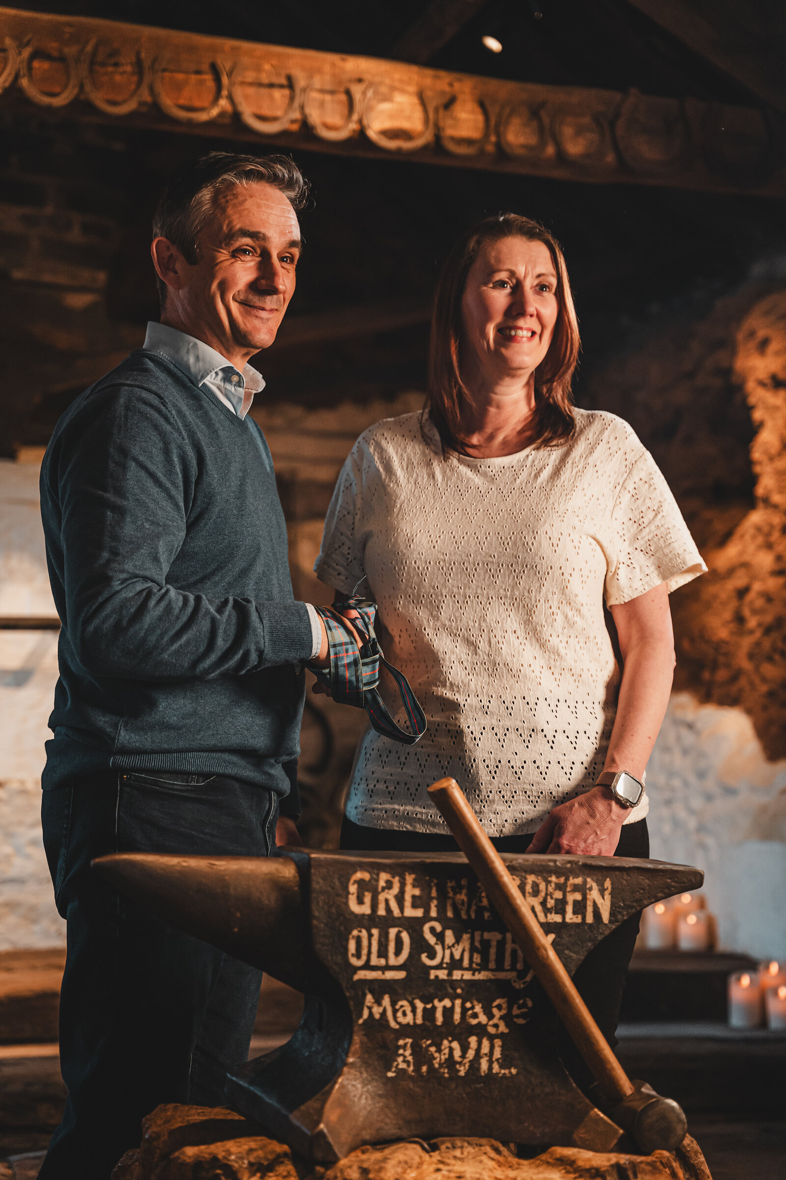 A couple stand smiling behind the anvil after their handfasting in the old smithy.
