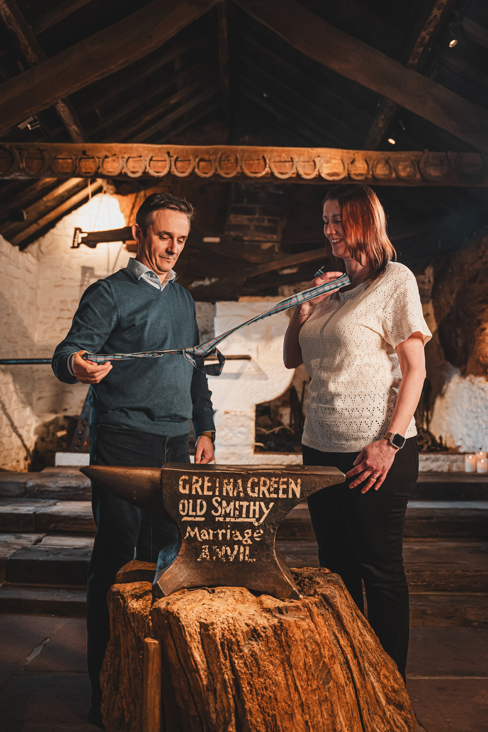 A couple hold the tartan ribbon over the marriage anvil in the low-beamed old smithy.