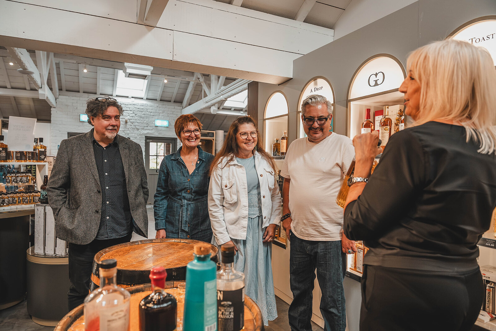A group listens to their host at the barrel table in the tasting room, shelves of bottles behind.