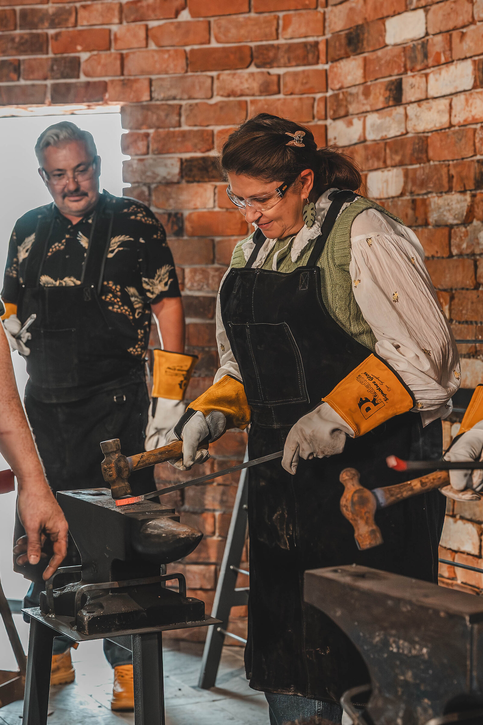 Two visitors in aprons and safety glasses hammer at their anvils side by side.