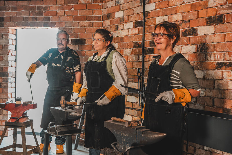 Three people in leather aprons and gloves work hot steel at their own anvils in the Gretna Green Forge.
