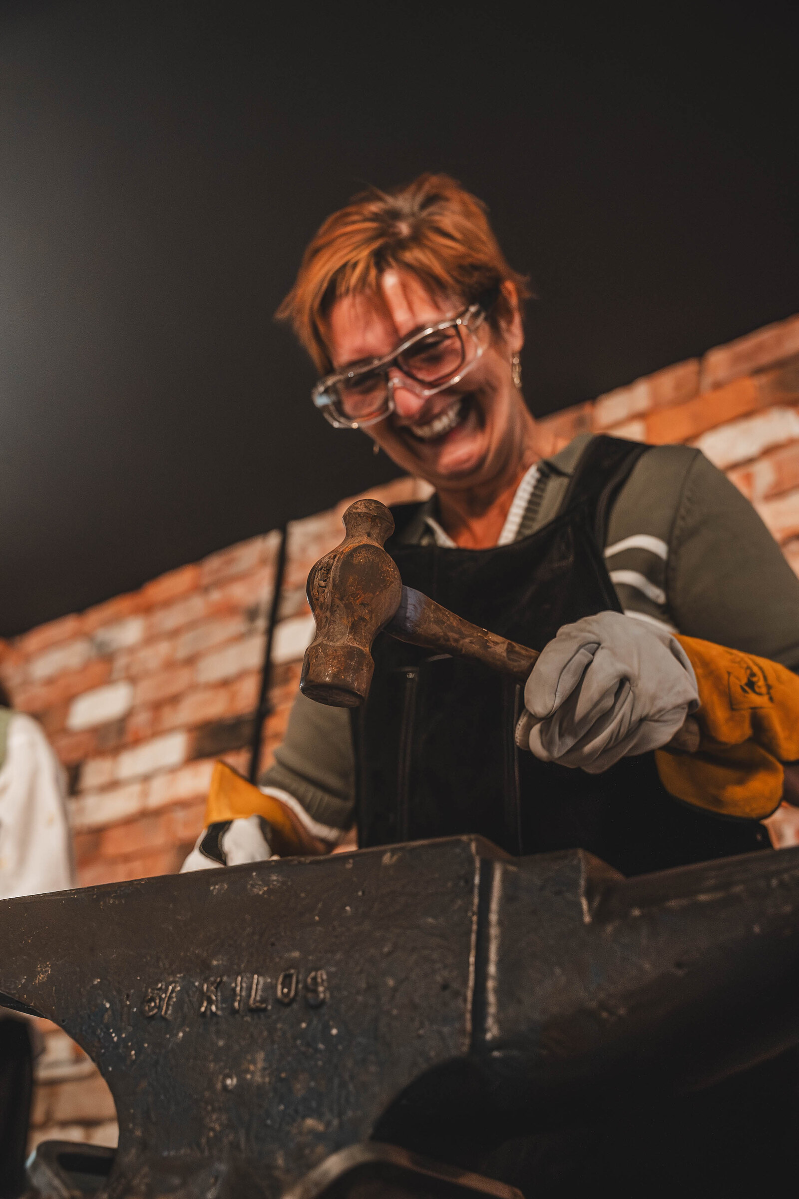 A woman in safety glasses laughs as she brings her hammer down on hot steel at the anvil.