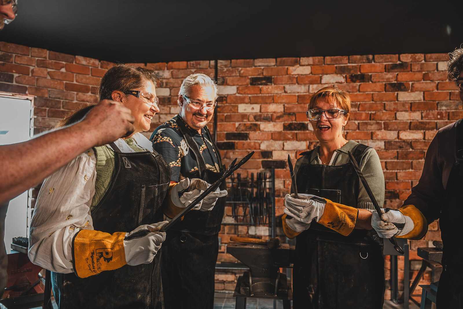 Visitors in aprons and gloves show off their work at the anvil, smiling in the Forge.