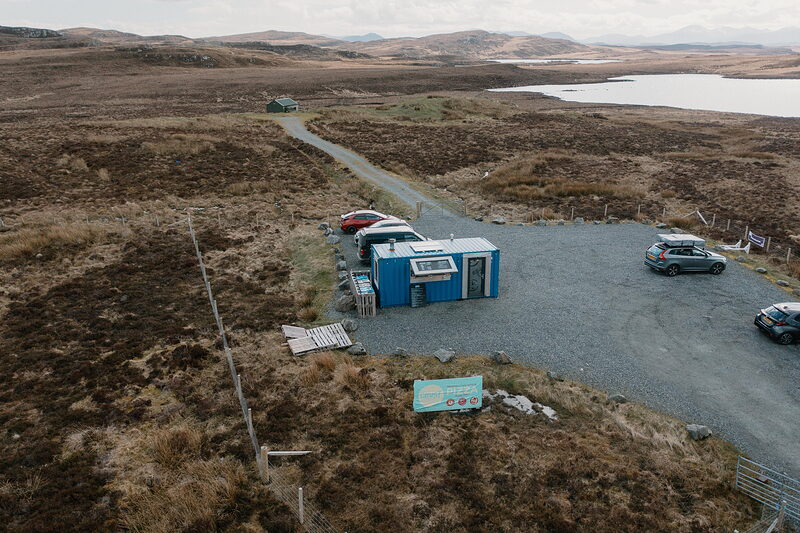 An aerial view of the blue Crust kiosk standing in a gravel car park beside a loch, cars parked around it and open moorland stretching behind.