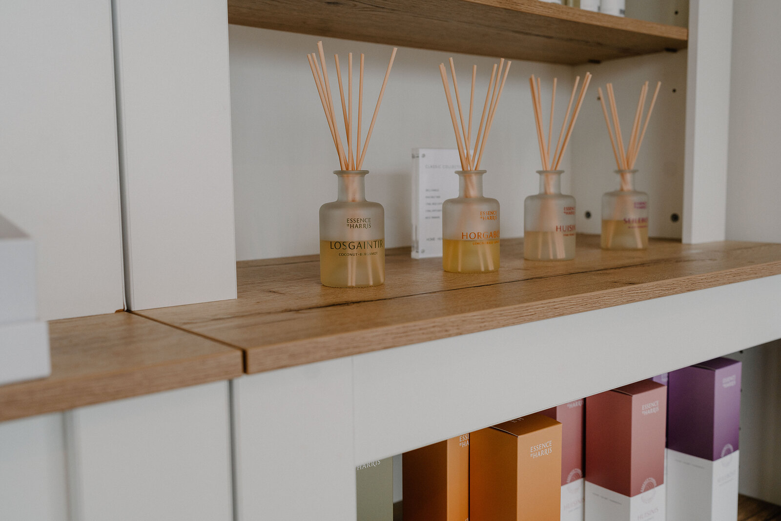 Four glass reed-diffuser bottles in a row along a wooden shelf edge, with retail gift boxes in graduated colours arranged beneath.